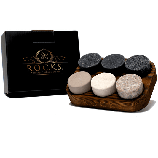 six whiskey stones are placed in their wooden container that has ROCKS engraved on the side, they are sitting next to a black packaging box with r.o.c.k.s. whiskey chilling stones in gold