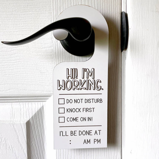 white dry erase finished door hanger is hanging on a door handle and says hi i'm working and has a check box next to do not disturb, knock first, and come on in!  beneath this it has a spot to insert a time where it says I'll be done at